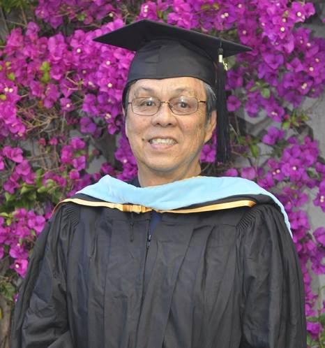 A woman in a graduation cap and gown is standing in front of purple flowers.