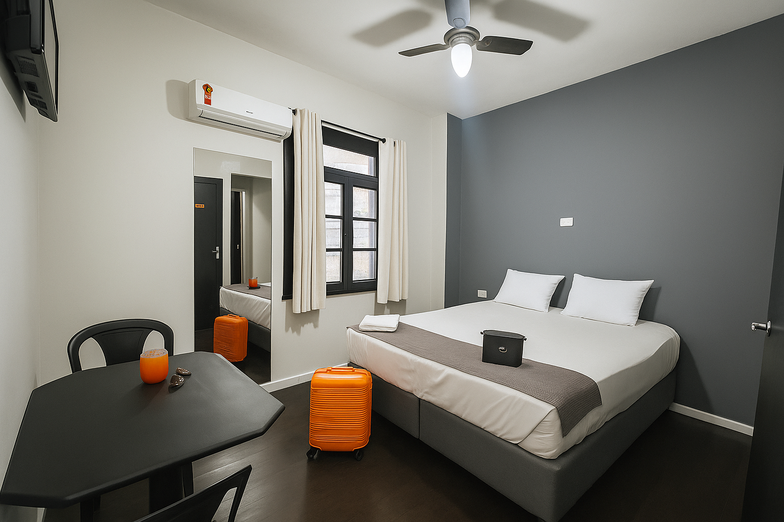 Quarto com cama, mesa, malas, espelho, parede cinza escuro e ventilador de teto.