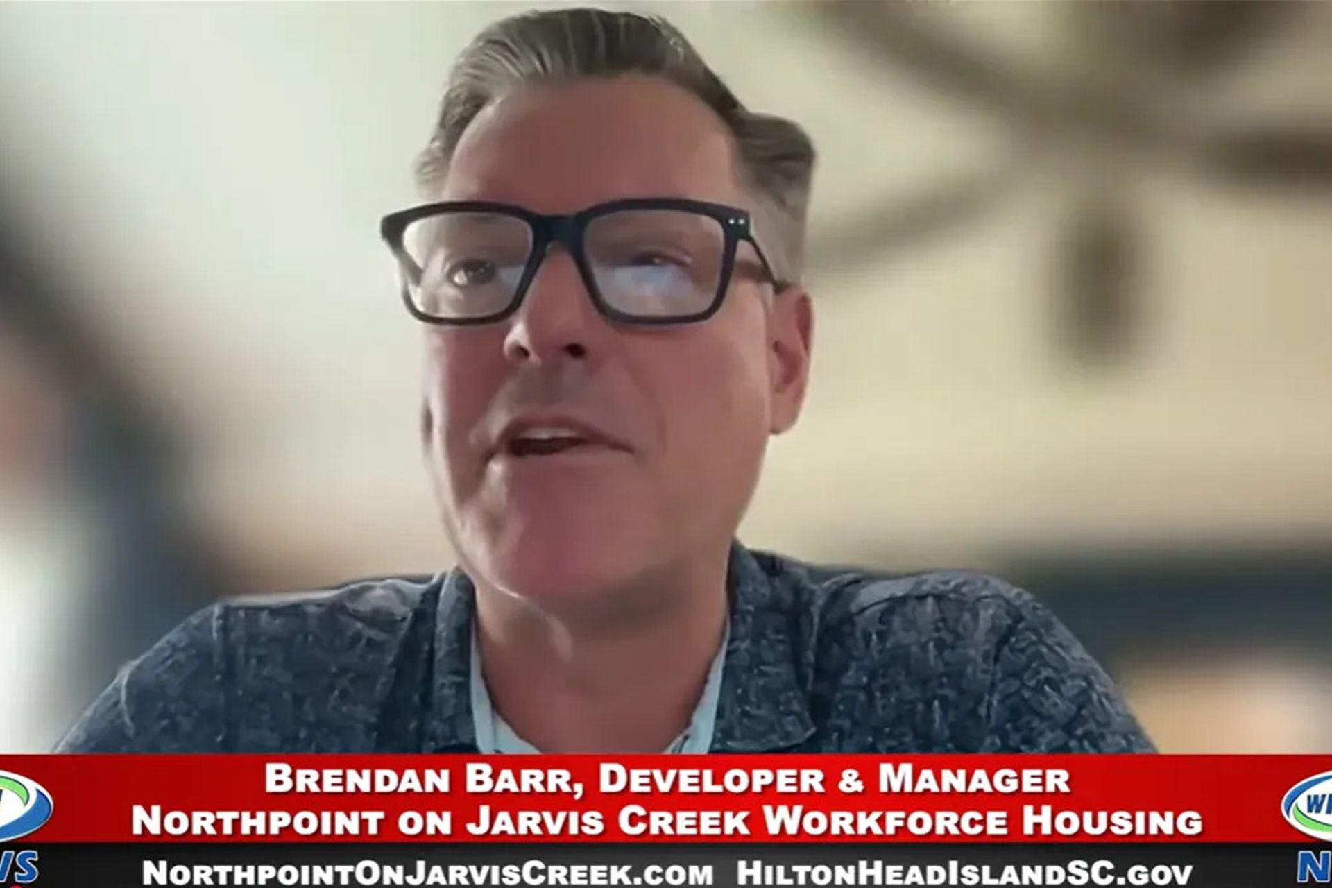 Man in glasses speaking on a video call, with a red lower-third banner and blurred ceiling fan background.
