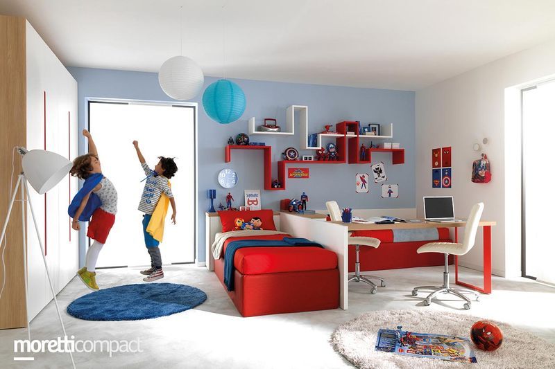 Cameretta colorata per bambini con due bambini vicino alla porta, letto rosso, mensole, scrivania e lanterne di carta appese.