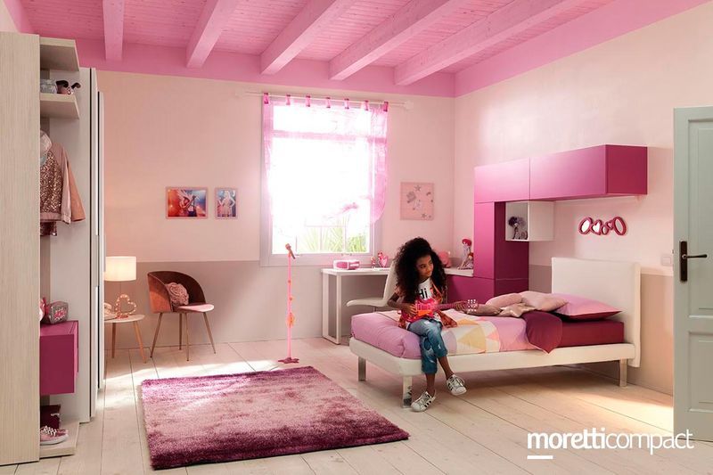 Camera da letto rosa con un bambino seduto su un letto, mobili bianchi e una finestra con tende trasparenti.