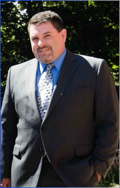 Man in a dark suit and blue shirt standing outdoors in sunlight with trees in the background