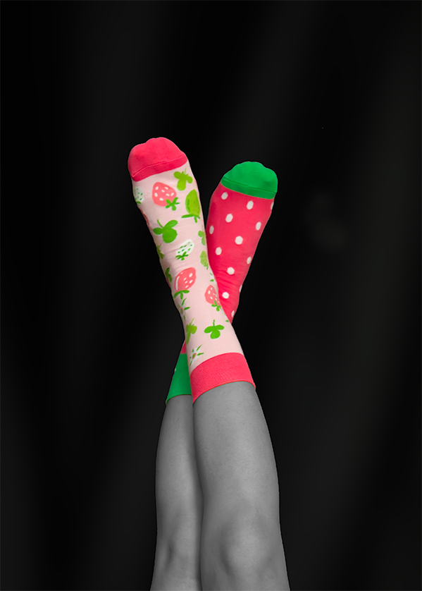 Crossed legs in colorful socks: one pink with strawberries, one red with white polka dots, set against a black backdrop.