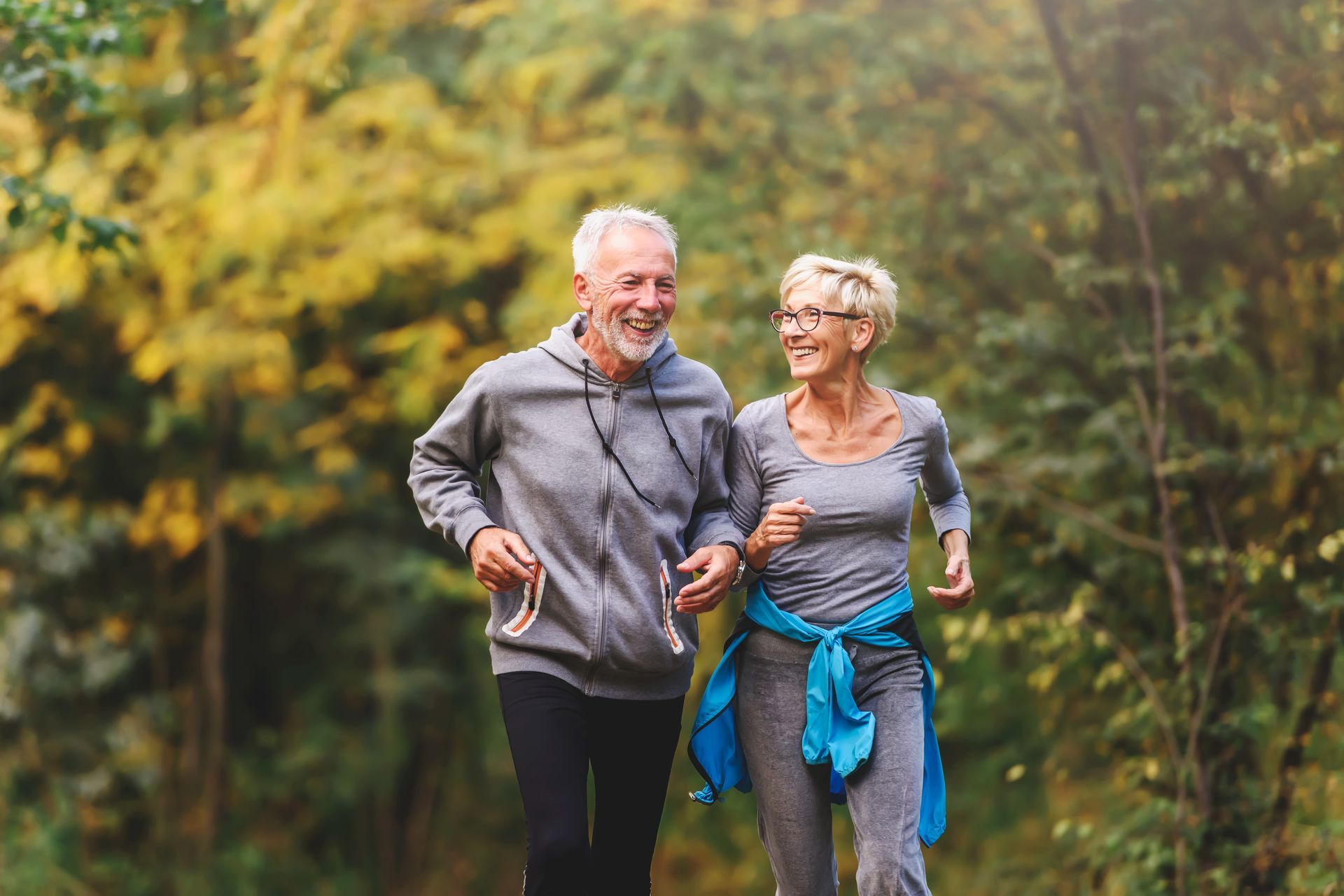 Two hikers smiling on a forest trail, one in gray with a blue jacket tied at the waist.