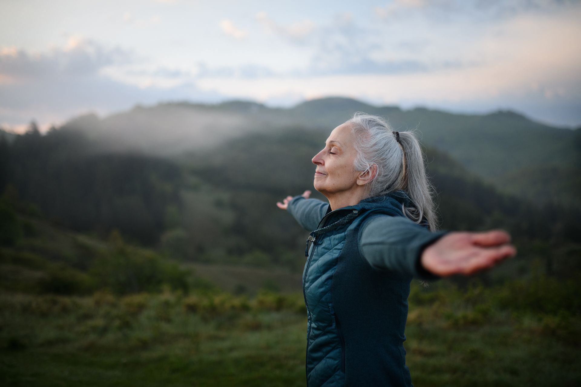 Woman in a teal jacket stands with arms outstretched in a misty mountain landscape at dawn Woman in a teal jacket stands with arms outstretched in a misty mountain landscape at dawn