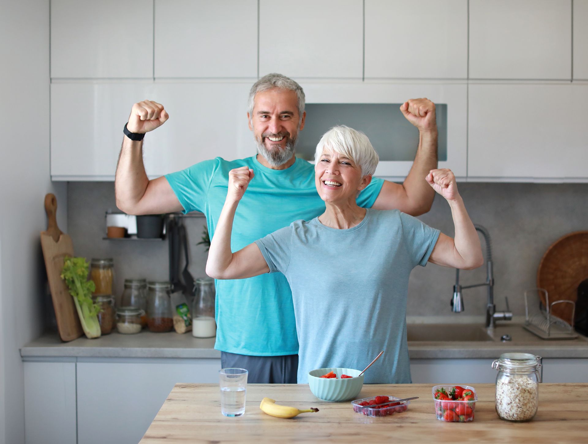 Two people flexing in a bright kitchen, smiling behind a counter with breakfast items. Two people flexing in a bright kitchen, smiling behind a counter with breakfast items.