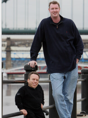 Two men of contrasting heights stand outdoors by a metal railing with a bridge in the background.