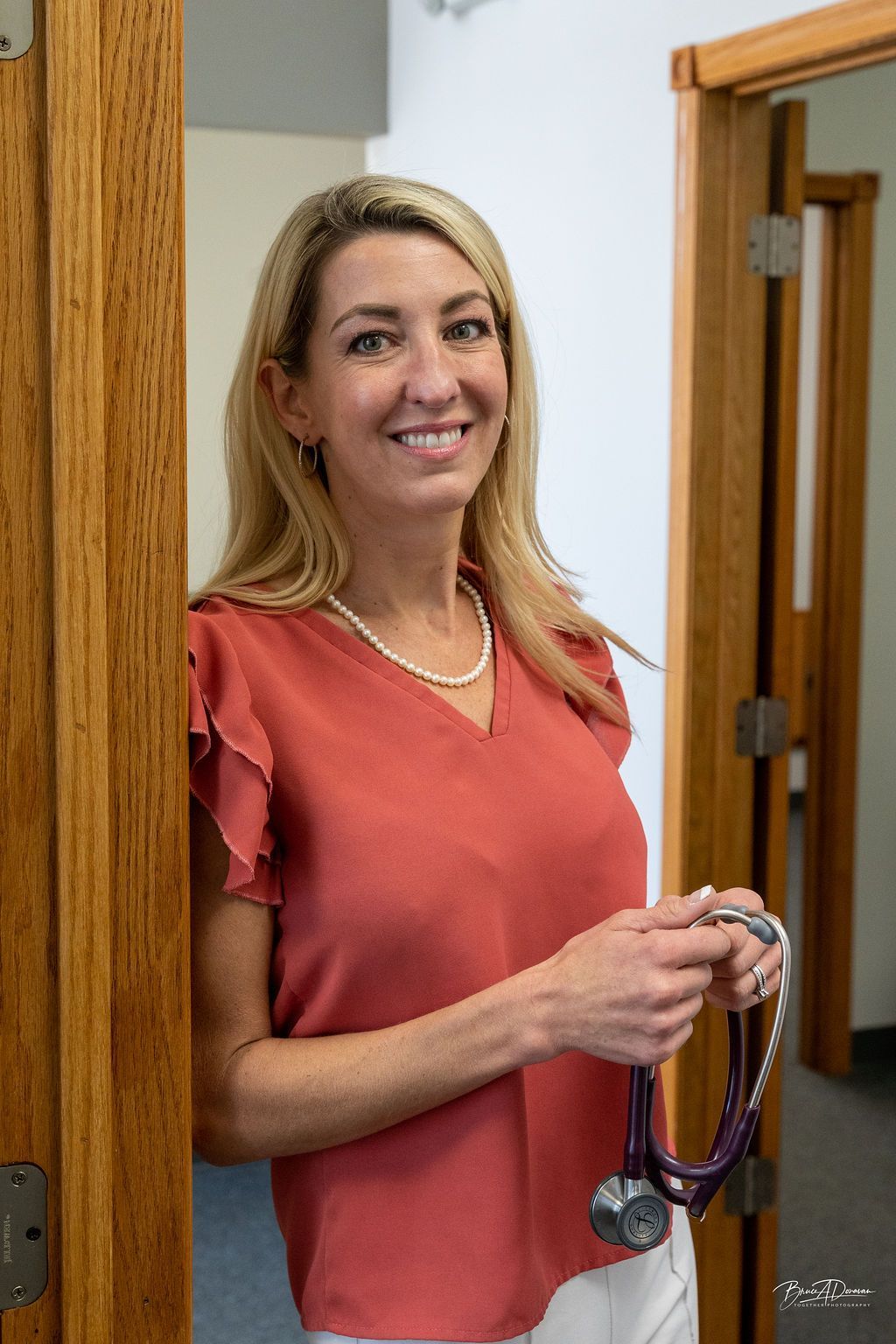Blonde woman, leaning in doorway, holding stethoscope, wearing red top, smiling.