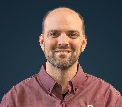 Man with a beard and a maroon button-down shirt smiles, standing in front of a blue background.