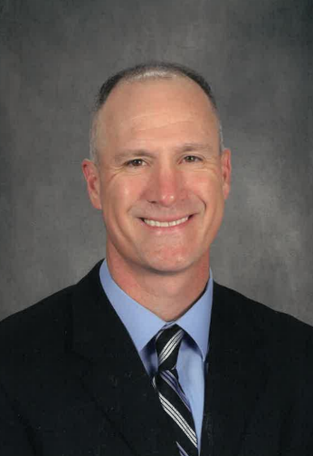 Man in suit smiles at the camera, light skin, blue shirt, striped tie, and dark background.