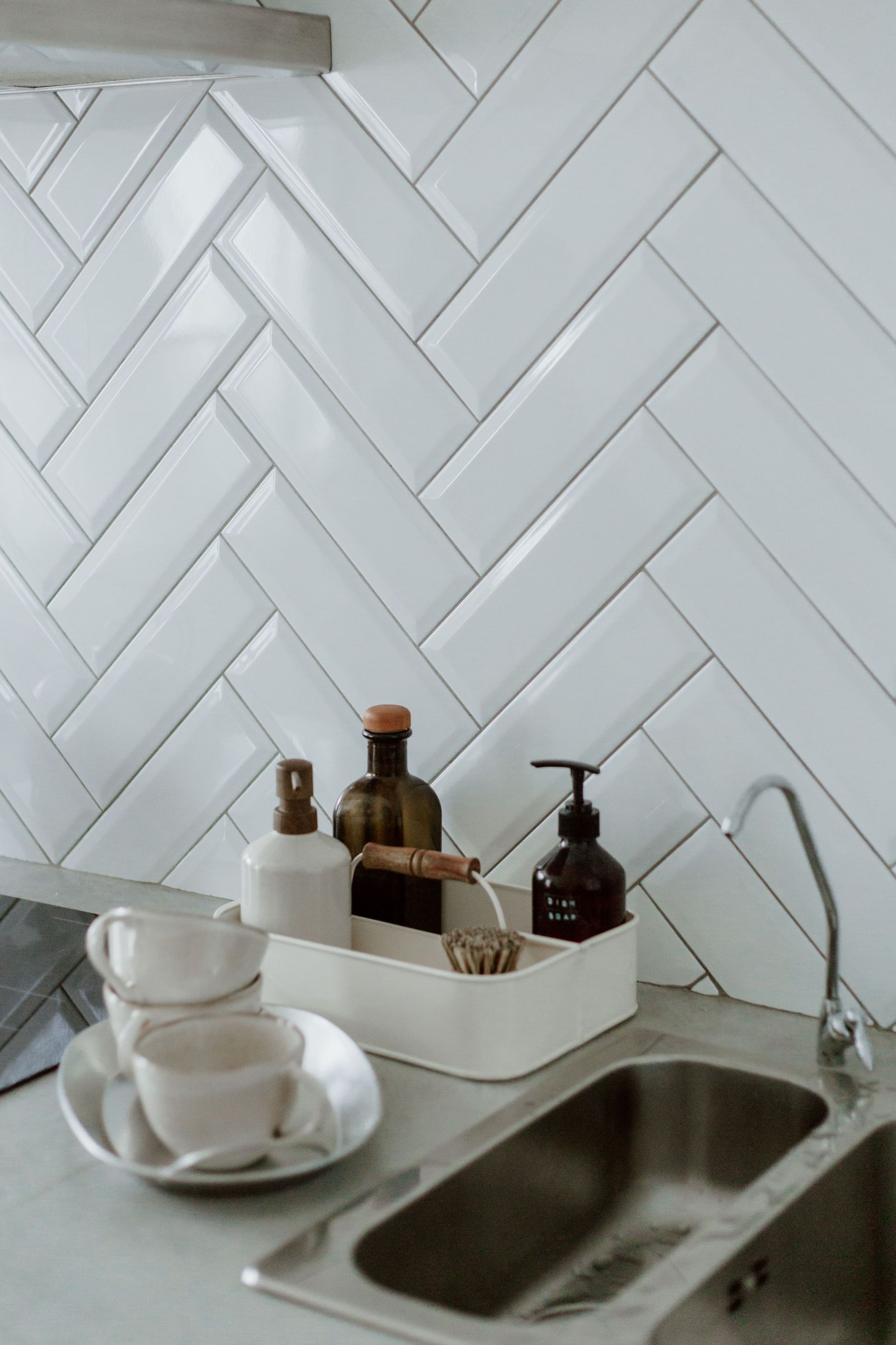 Kitchen sink with white herringbone tile backsplash, dish soap, cups, and a faucet
