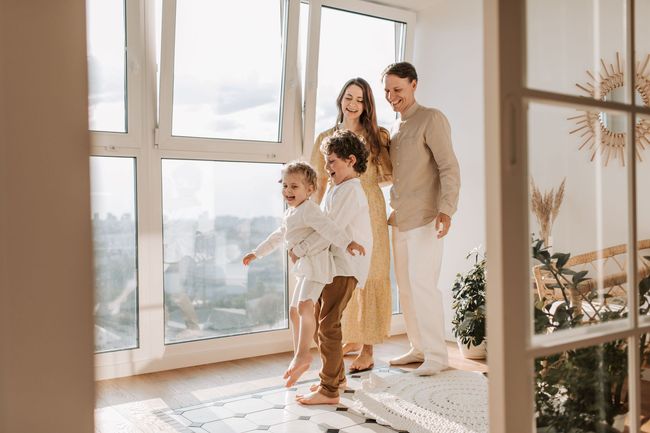 Family standing by bright floor-to-ceiling windows in a sunlit living room