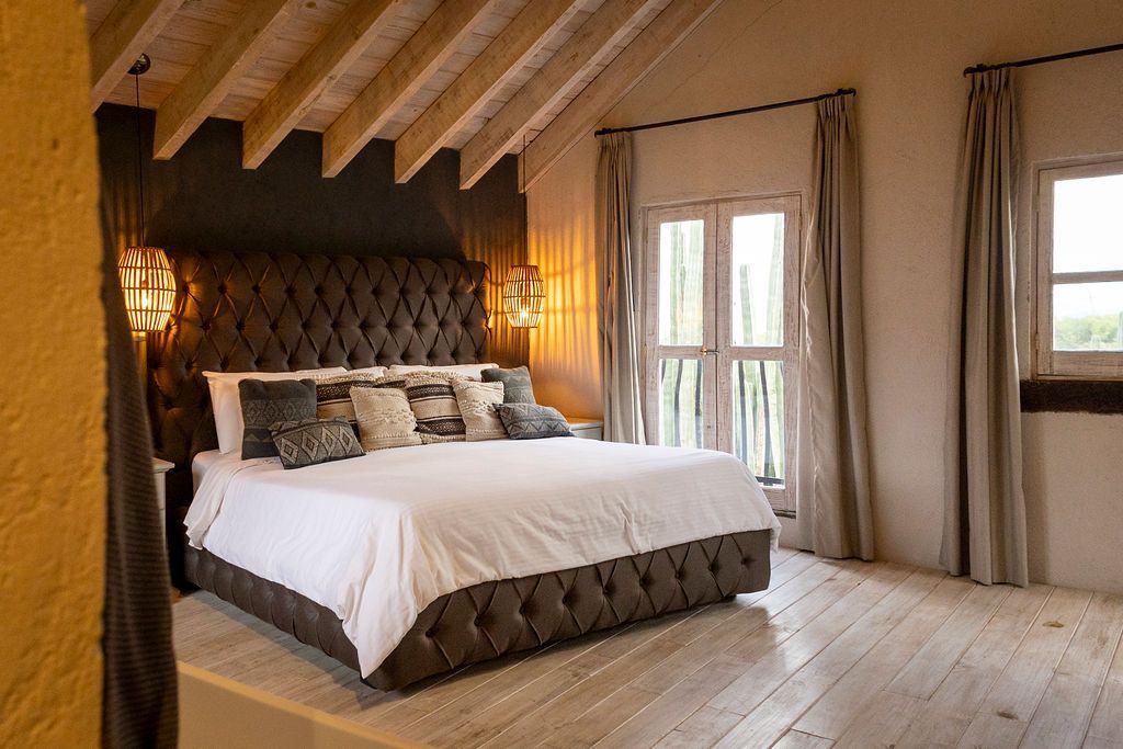 Bedroom with a large bed, tufted headboard, and two windows with light curtains.