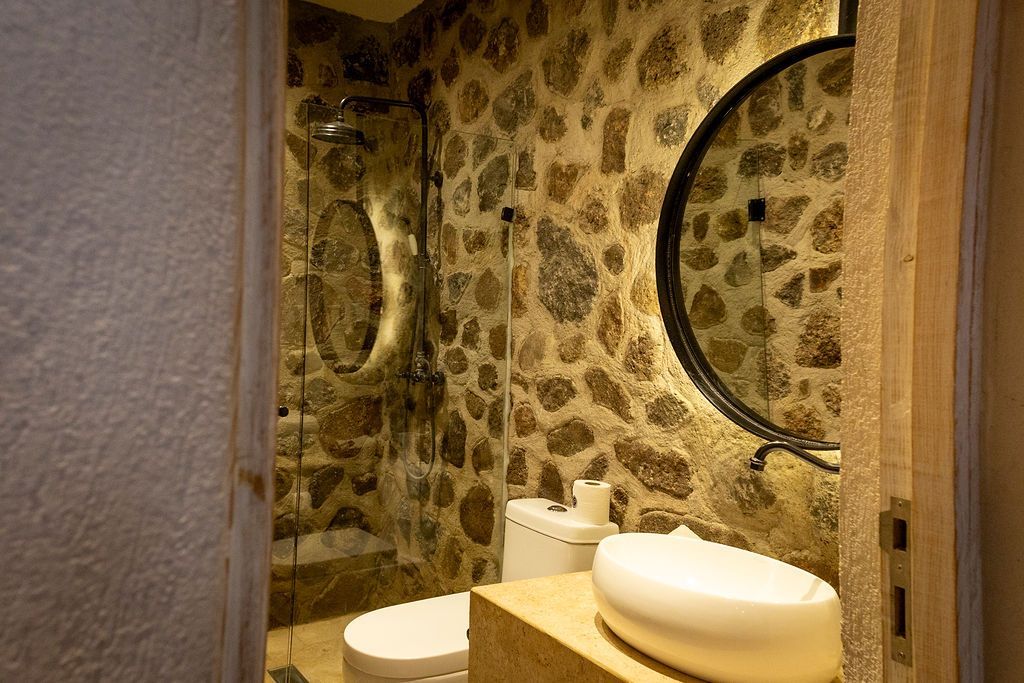 Bathroom with stone wall, round mirror, white sink and toilet.