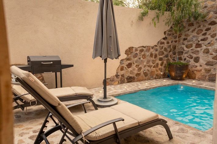 Small pool area with lounge chairs, umbrella, and grill. Stone wall and blue water.