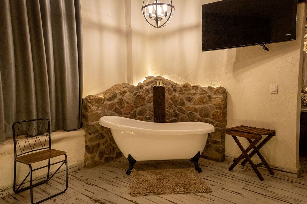 Claw-foot tub in a rustic bathroom with stone wall, chair, small wooden table, and mounted TV.
