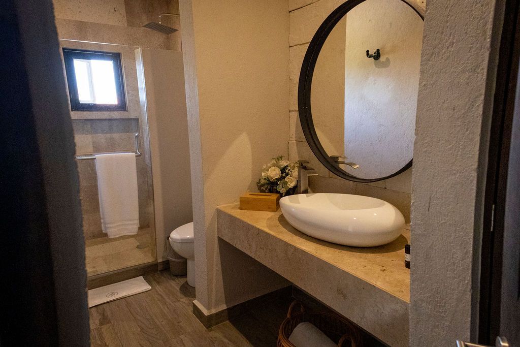 Bathroom with stone walls, wooden floor, oval sink, and a round mirror. A shower and toilet are also visible.