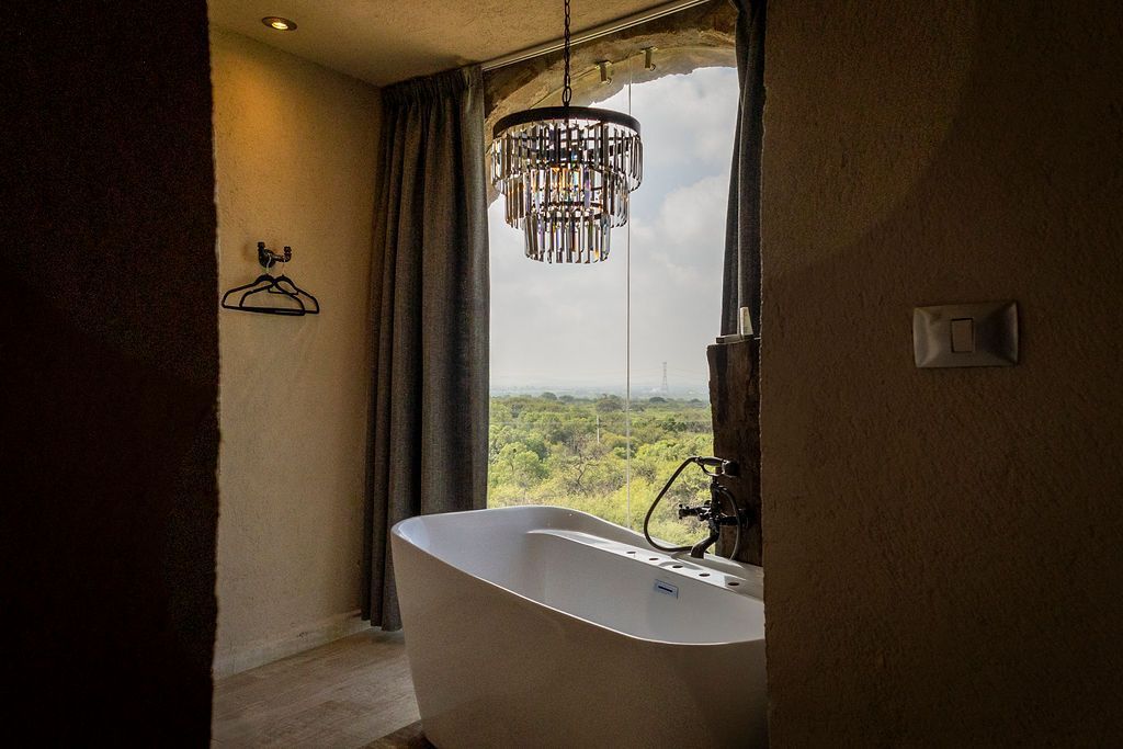 Bathroom with freestanding tub, chandelier, large window with forest view, grey curtains.