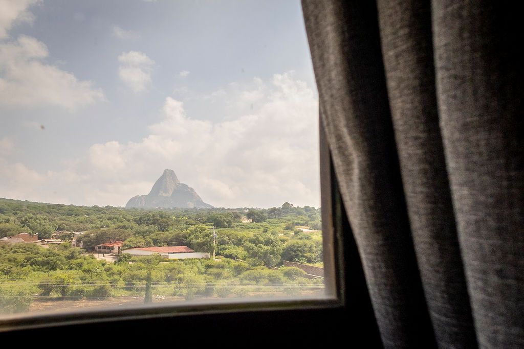 View from window with curtain, mountain in the distance over green trees, and houses.