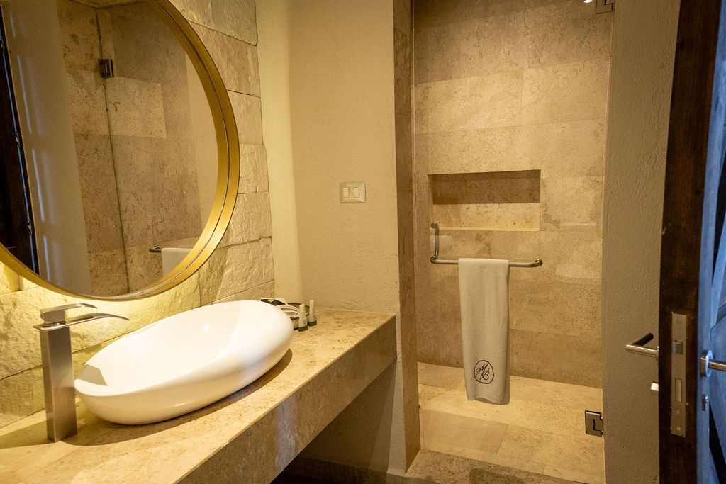 Bathroom with beige stone walls, round mirror, white sink, and shower.