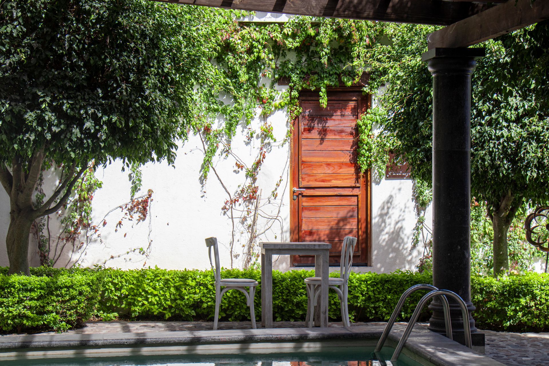 Outdoor patio with pool. White wall with wooden door, table, two chairs, and lush greenery.