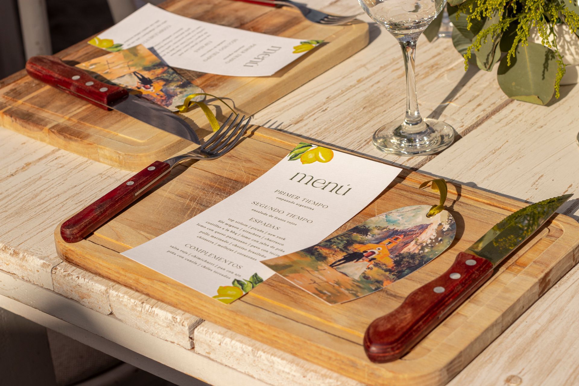 Two wooden boards with menus, knives, and forks on a white table set for a meal.
