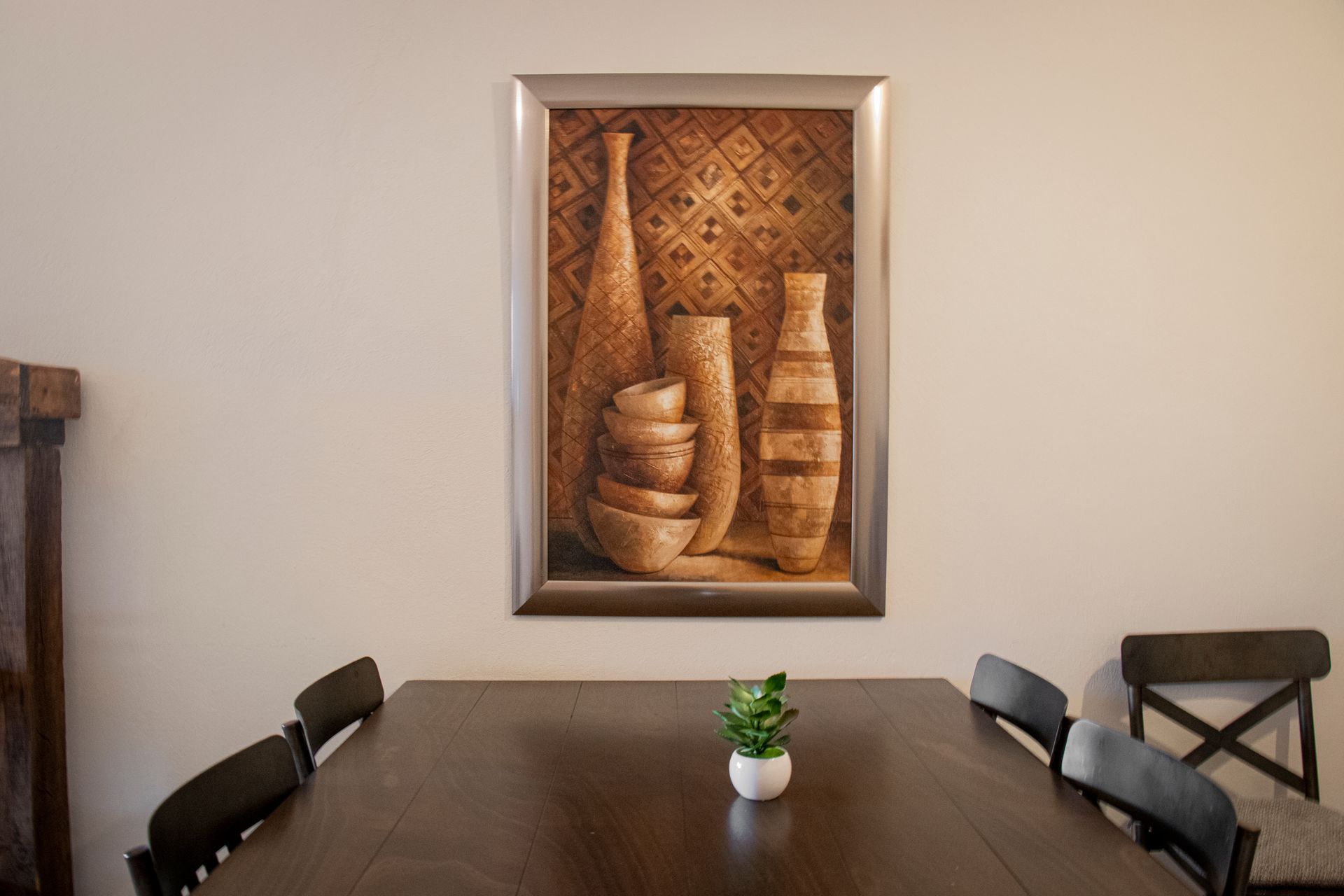 Dining room with art and a table set with a plant and chairs. Beige walls, brown wood.
