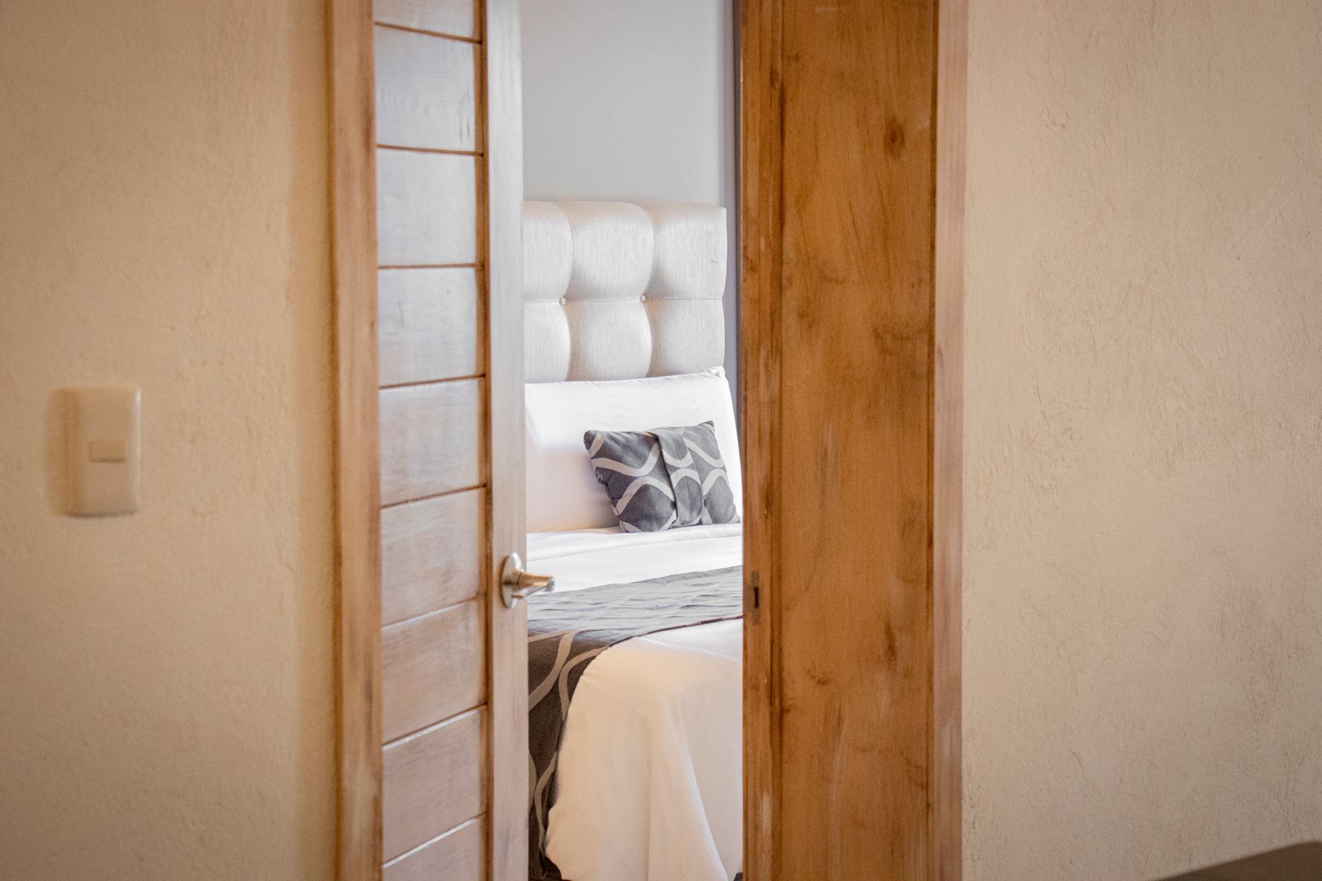 Open wooden door leading into a bedroom with a bed, light-colored walls, and a tufted headboard.