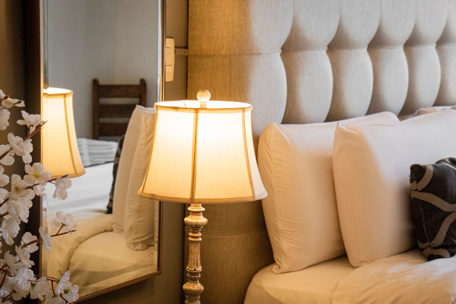 Beige tufted headboard with a lit lamp on a stand beside the bed, reflected in a large mirror.