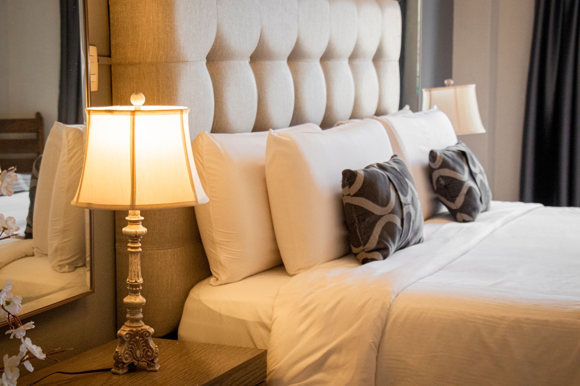 Cozy bedroom with a tufted headboard, crisp white bedding, and decorative pillows. A lamp sits on the bedside table.
