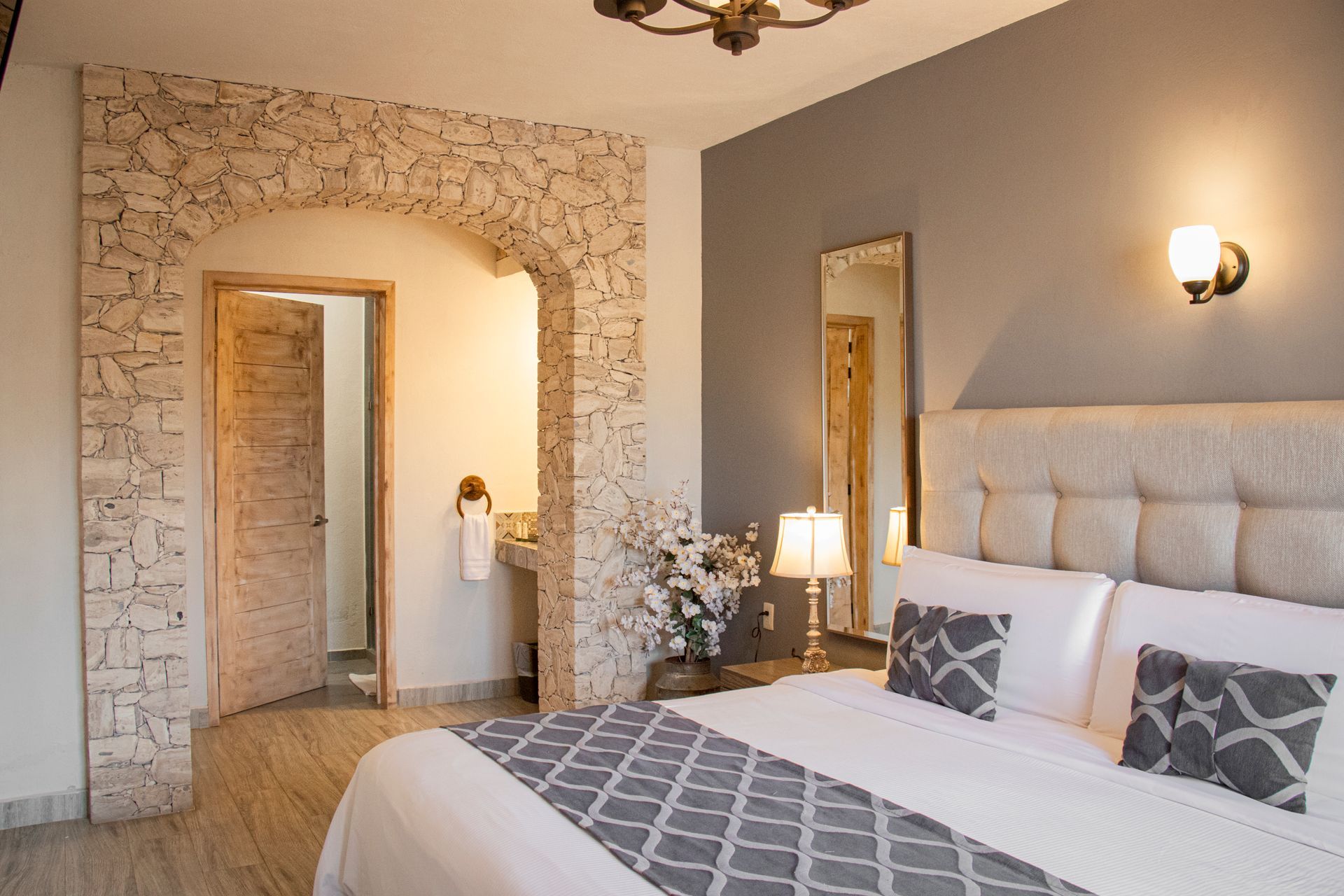 Bedroom with arched stone doorway to a bathroom. Beige and gray color scheme, a bed with decorative pillows.