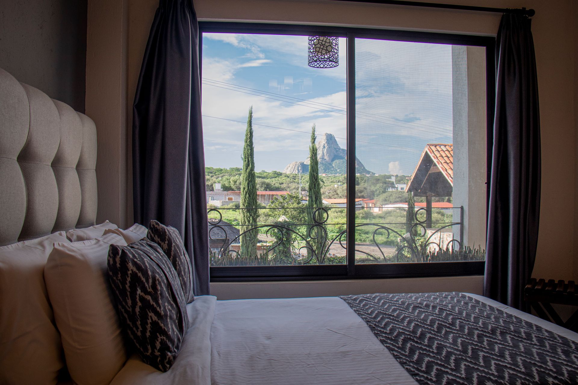 Bedroom with view: white bed, gray headboard, large window framing outdoor scenery of trees and buildings.