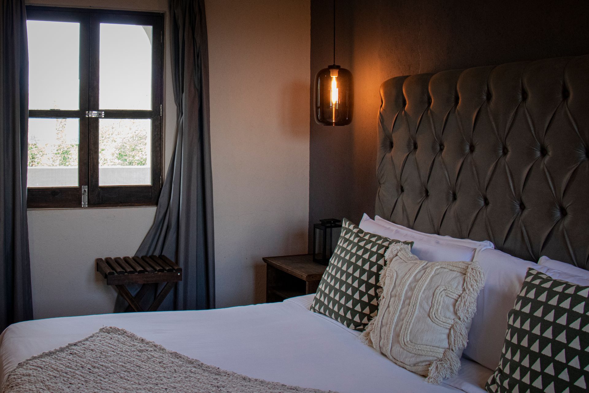 Bedroom with tufted headboard, window, bed, decorative pillows, and hanging light.