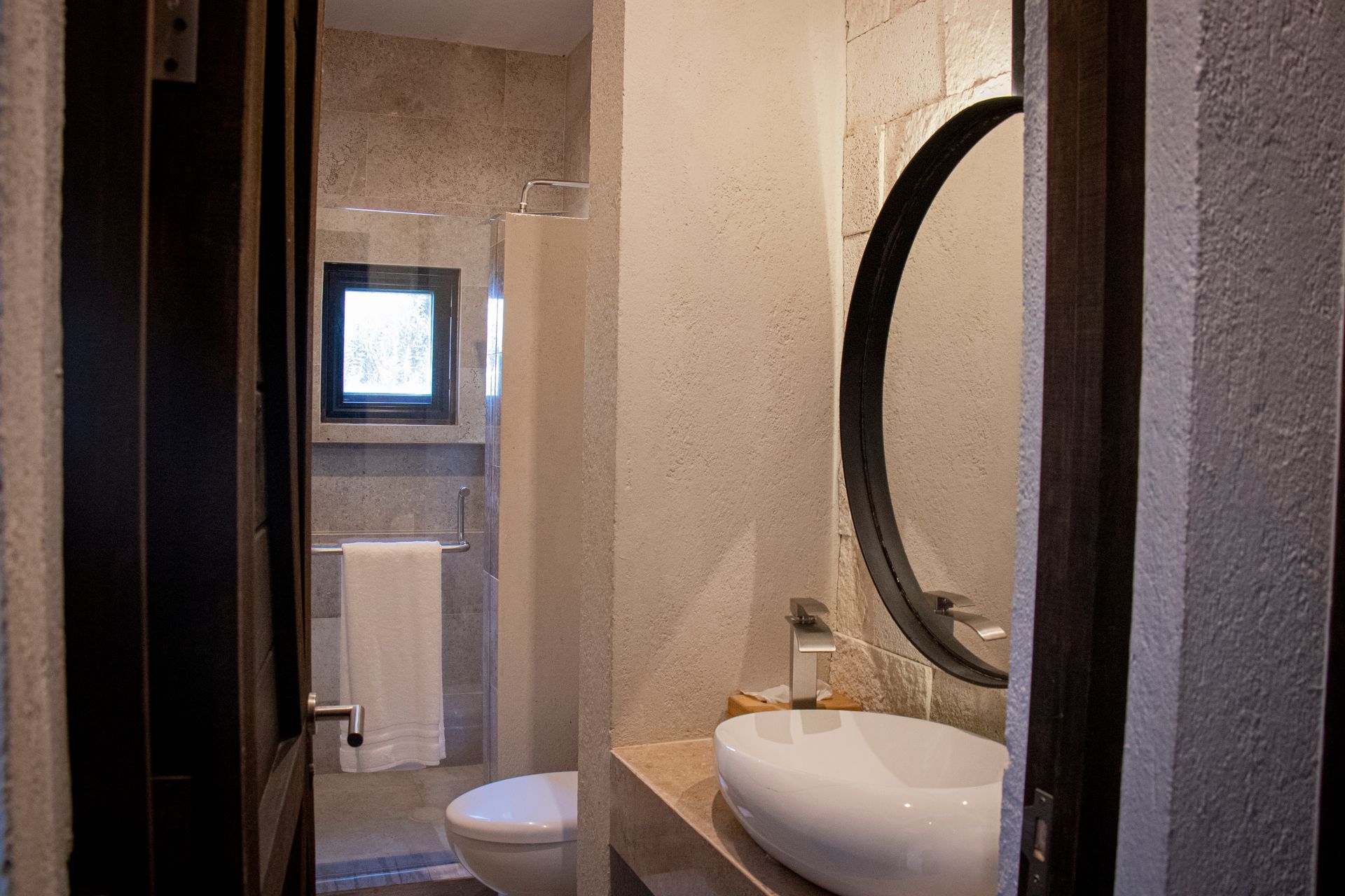 Bathroom interior: white sink, circular mirror, toilet, partially visible shower, beige walls.