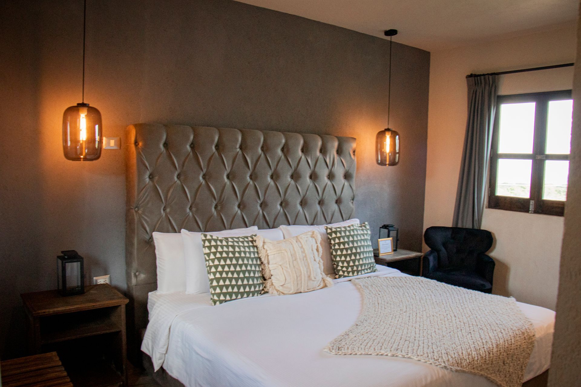 Bedroom with a tufted headboard, king-size bed, side tables, and pendant lights. A dark chair sits beside a window.