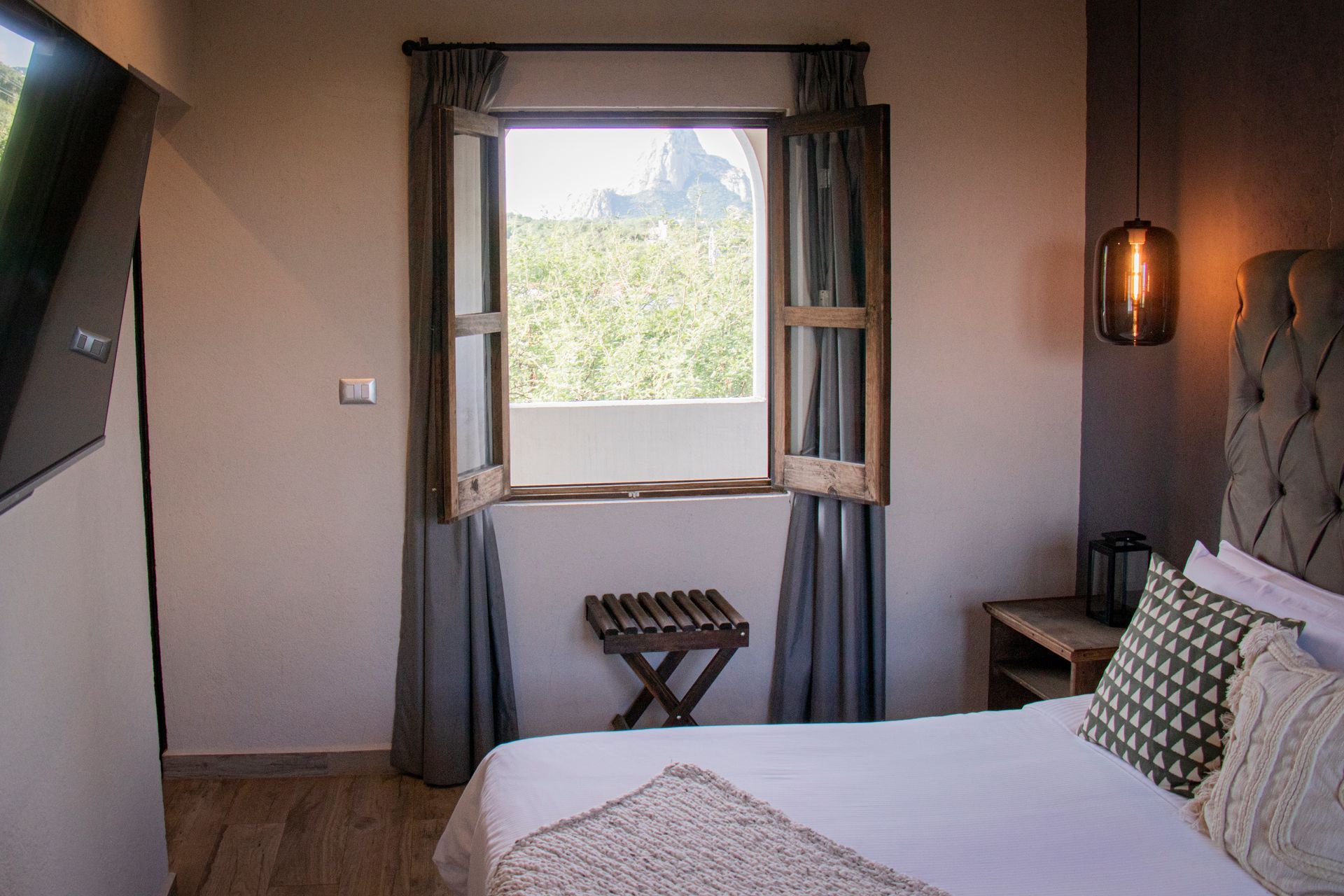 Bedroom with an open window, curtains, a bed, a small stool, and a hanging light.