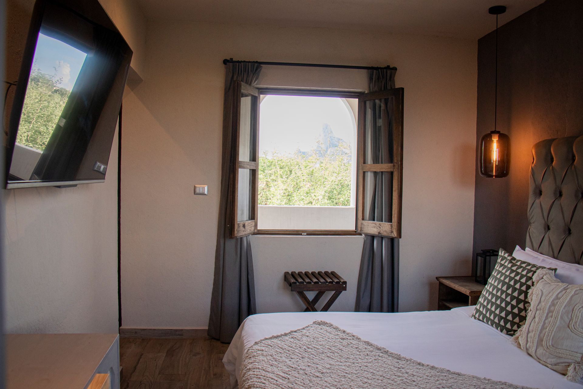 Bedroom with bed, window, TV, and decorative elements. Light walls and wooden floor.