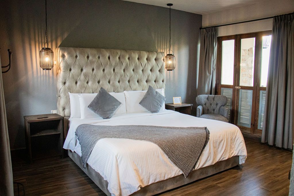 Bedroom with a large tufted headboard, king-sized bed, two hanging lights, a wooden floor, and a door to a balcony.