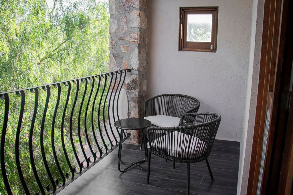 Balcony with black wrought-iron railing, two chairs, small table, and a view of green trees.