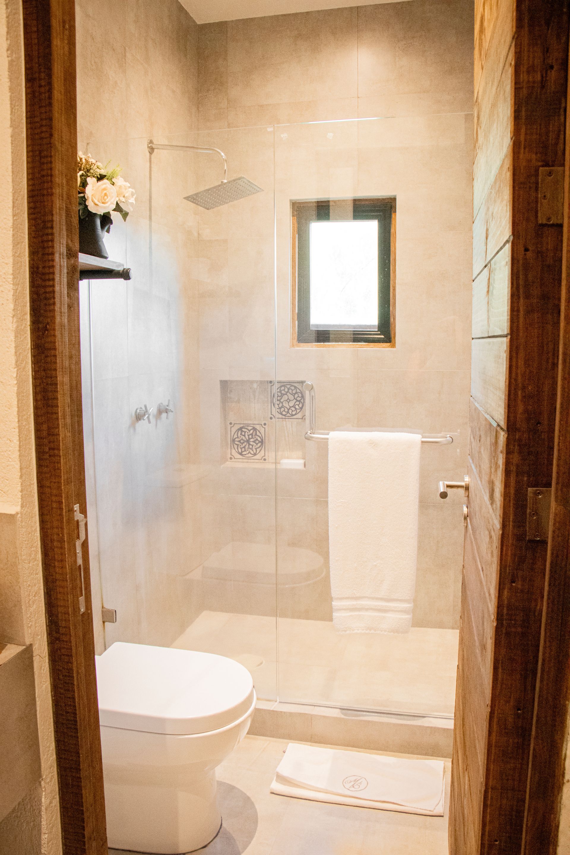 Bathroom with glass shower, toilet, and towel rack. Light gray walls and wooden door frame.