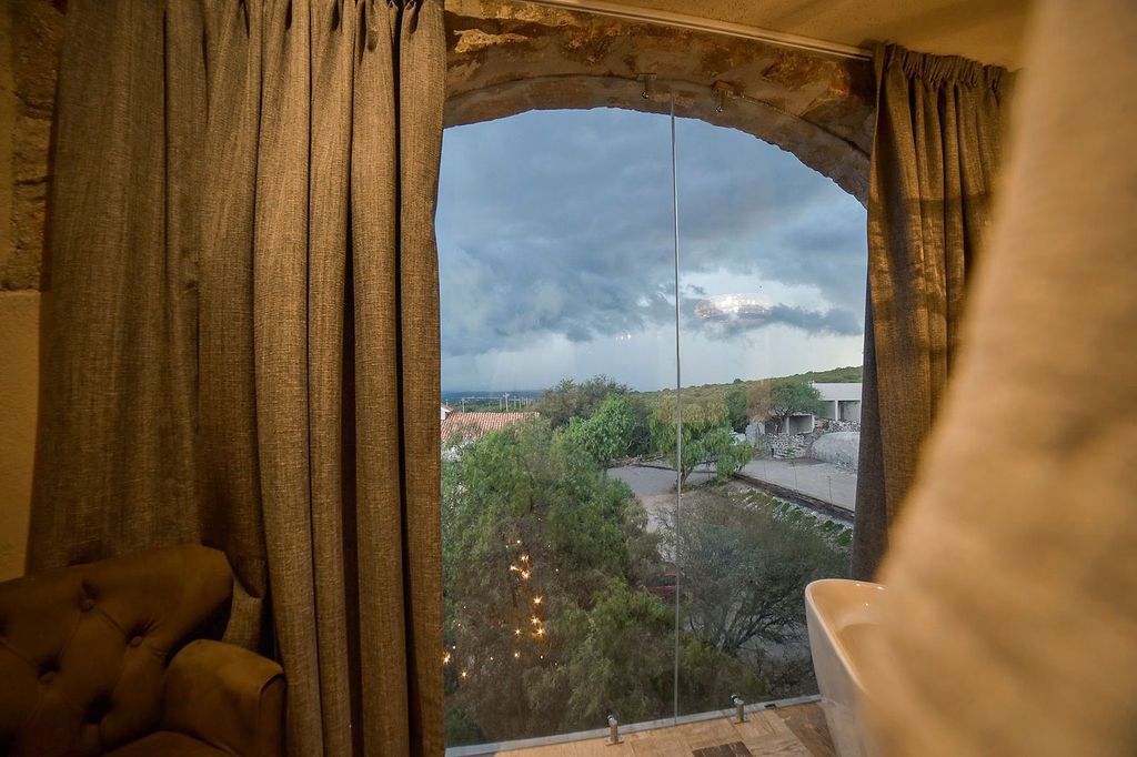 View through arched window: storm clouds, trees, and buildings. Curtains frame the view with an armchair on the left.