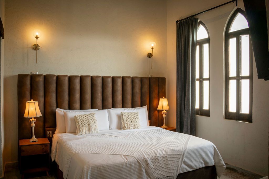 Bedroom with king-sized bed, tall headboard, nightstands, lamps, and arched windows. Soft lighting, neutral tones.