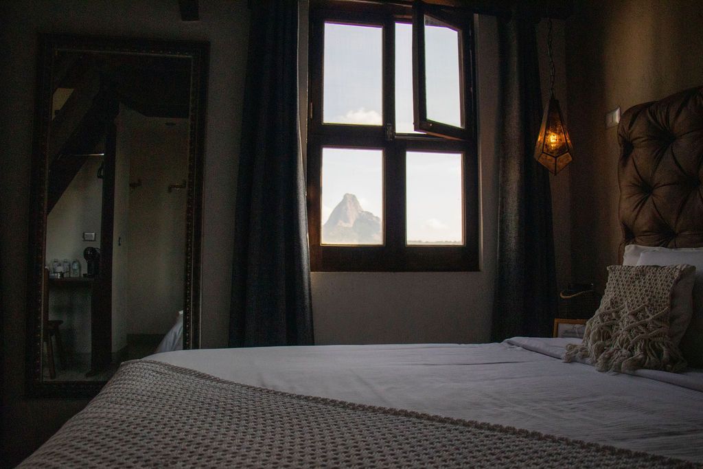 Bedroom with a window overlooking a mountain. Bed, mirror, and dark curtains.