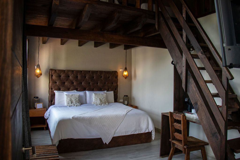 Bedroom with loft, featuring a bed, wooden desk and stairs. Dark wood beams and a brown tufted headboard.