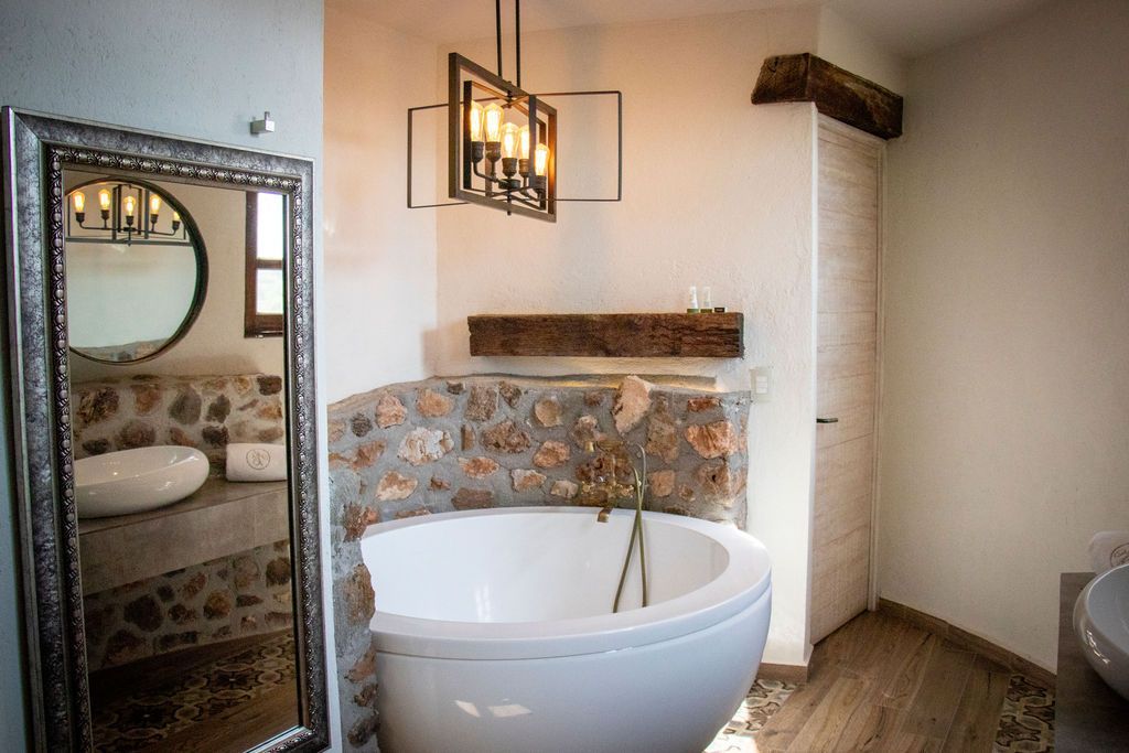 Bathroom with a white tub, stone wall, large mirror, and modern chandelier.