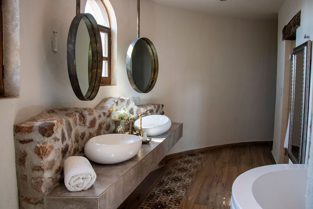 Bathroom with stone wall, two oval mirrors, white sinks, and wooden floor.