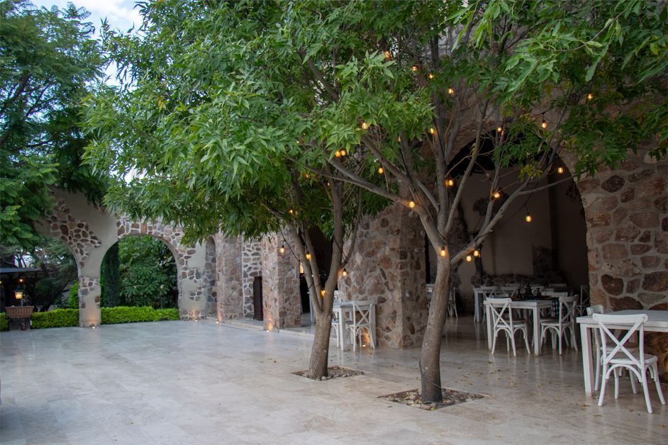 Outdoor event space with stone arches, trees with string lights, white tables and chairs.