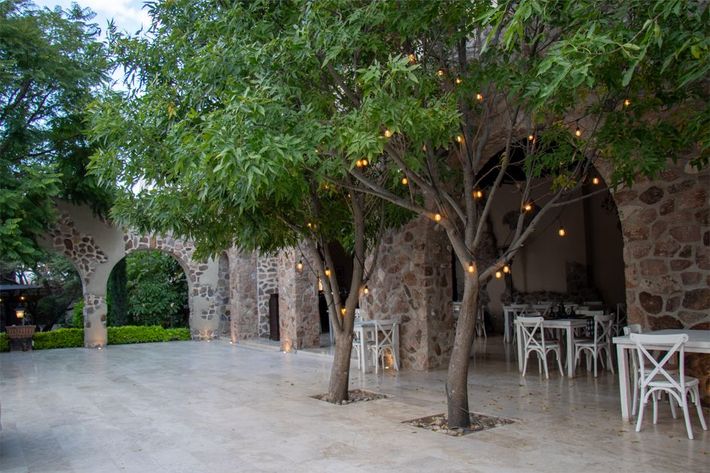 Outdoor event space with stone arches, trees with string lights, white tables and chairs.