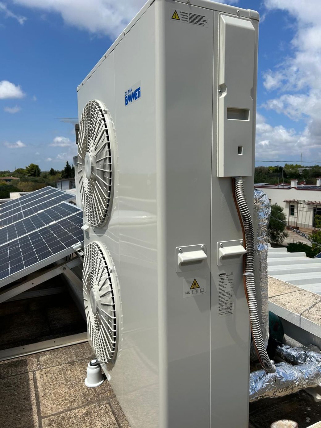 Unità esterna di climatizzazione posizionata accanto a pannelli solari su un tetto, con due grandi ventilatori e un mobiletto in metallo bianco.