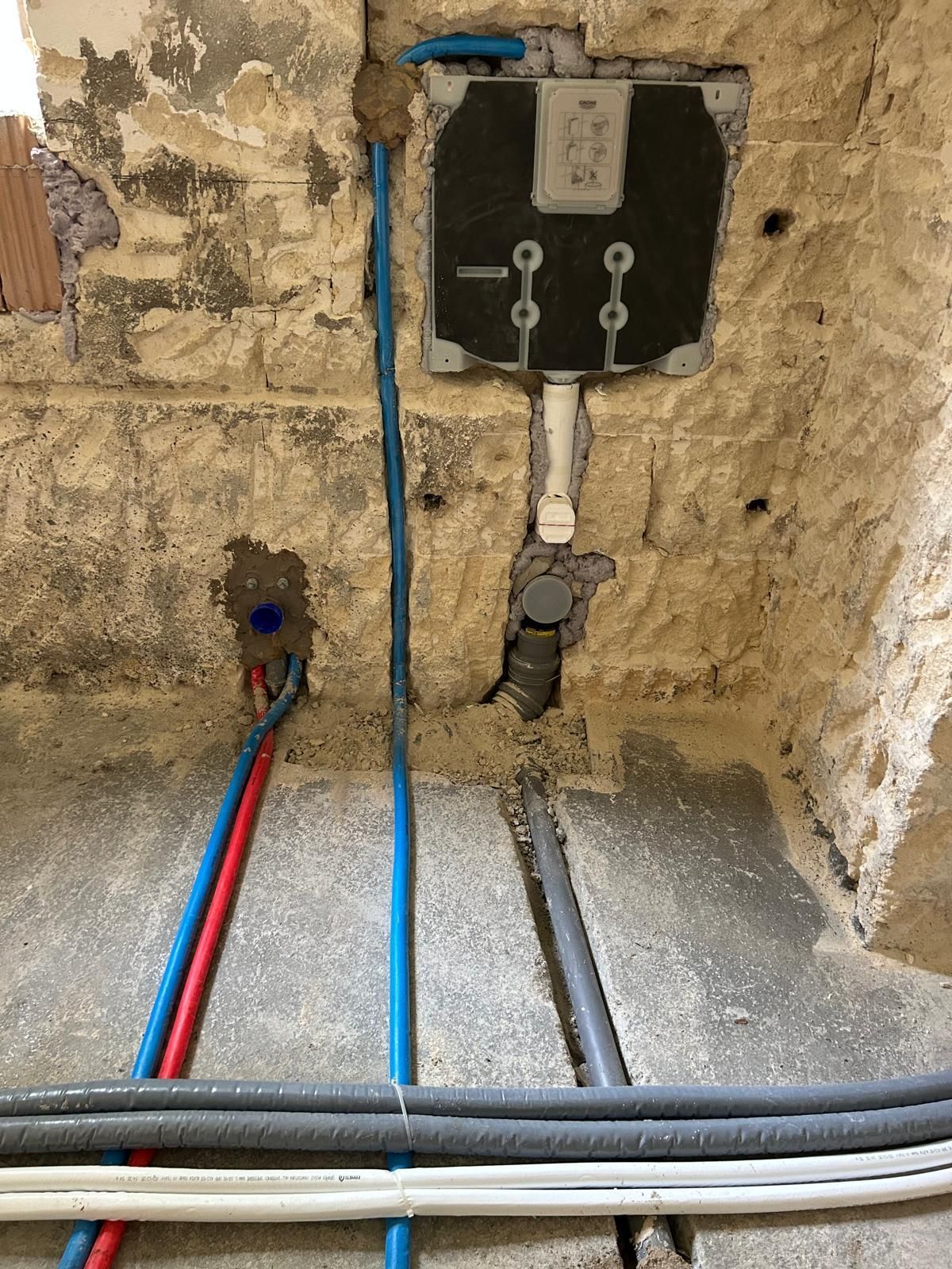 Tubature e cavi di servizio a vista su una parete grezza, con una scatola di controllo nera e linee blu e rosse sul pavimento.